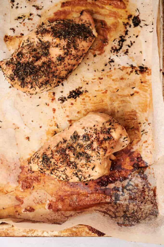 Two seasoned and baked chicken breasts rest on a parchment-lined baking sheet. The surface is slightly charred, with visible herbs, making them a perfect protein addition to a hearty Seven Layer Salad.