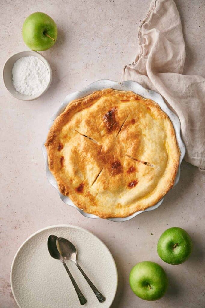 A baked pie with a golden crust on a white pie dish, accompanied by a bowl of powdered sugar, three green apples, a beige cloth, and two spoons on a plate, all set on a light-colored surface.
