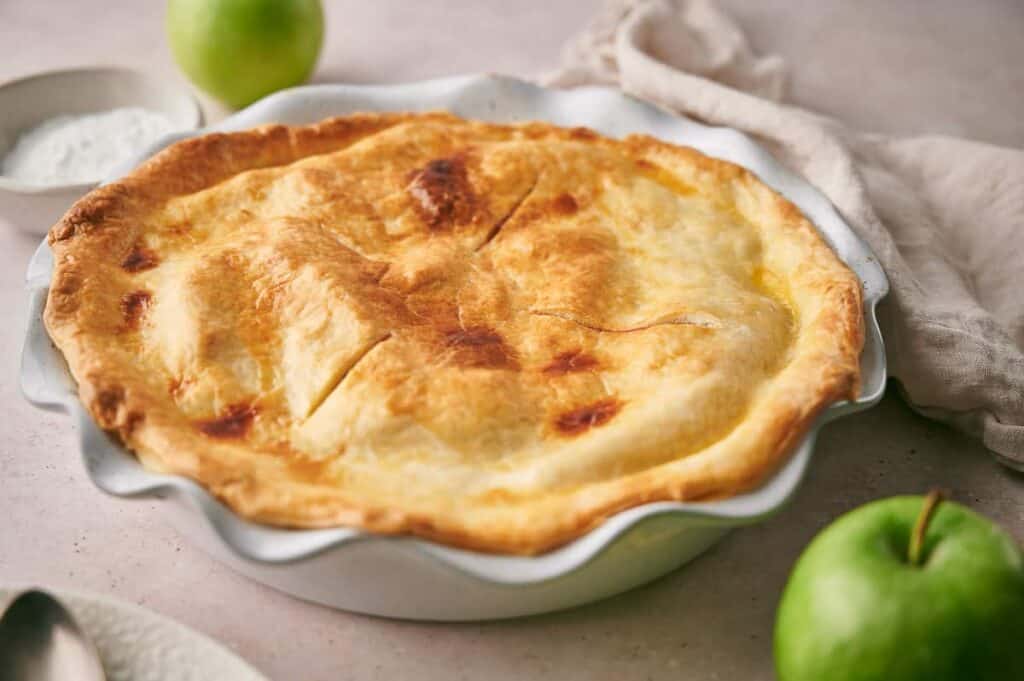 A freshly baked apple pie in a white ceramic pie dish, surrounded by green apples and a beige cloth.