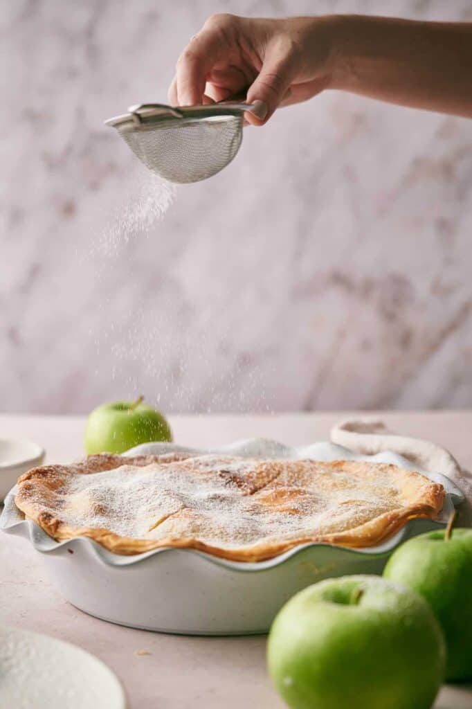 A hand sprinkles powdered sugar over a freshly baked pie in a fluted dish, with a few green apples on the table in the background.