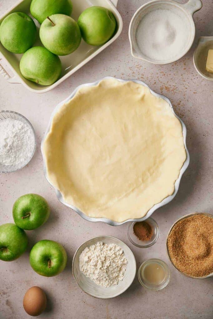A pie crust in a dish surrounded by green apples, sugar, butter, flour, an egg, and various baking ingredients on a light surface.
