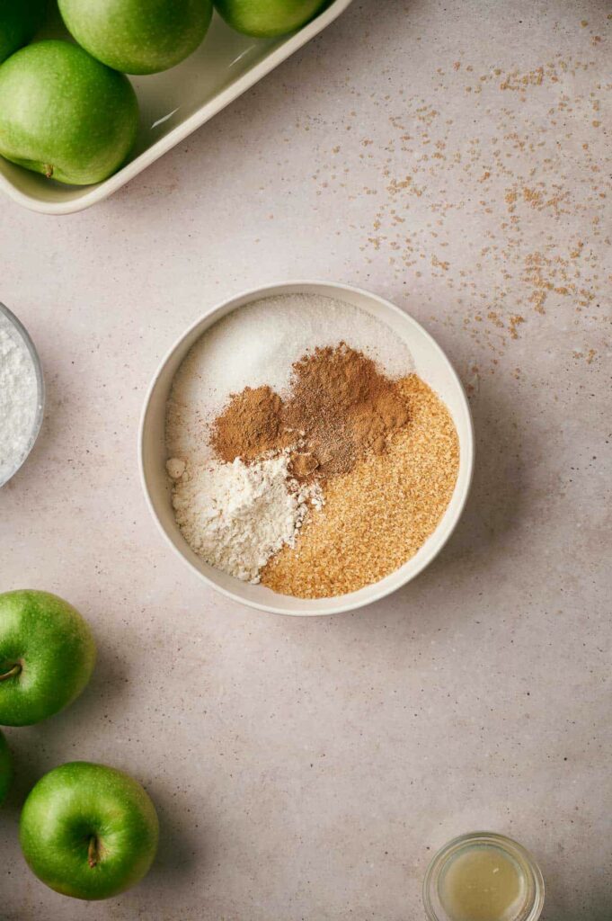 A bowl contains flour, sugar, cinnamon, and brown sugar, surrounded by green apples and a slice of a lemon on a light surface.