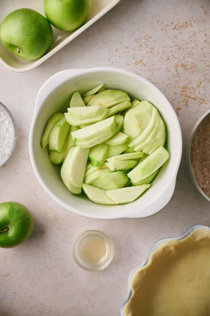 Top view of a bowl filled with sliced green apples, surrounded by whole green apples, a pie crust, a small dish of liquid, and a bowl of sugar on a light countertop.