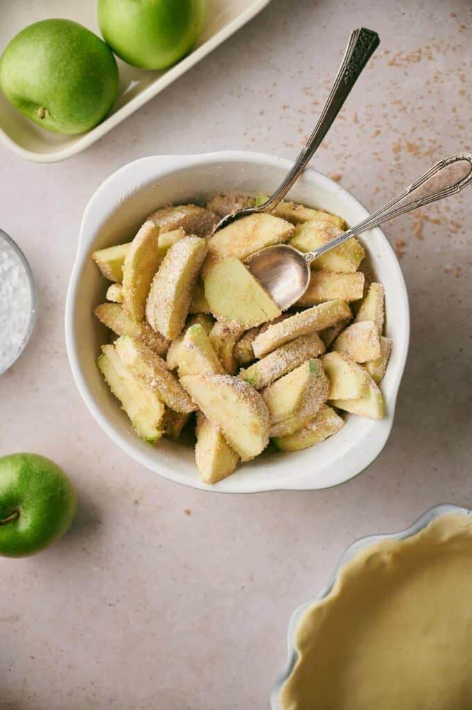 A bowl of sliced green apples coated with sugar and spices, with two spoons in it. Whole green apples, a white dish, and a pie crust are also visible on the table.