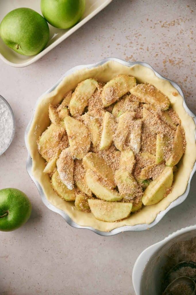 A pie crust filled with apple slices covered in a sugar mixture, next to green apples and baking ingredients on a countertop.