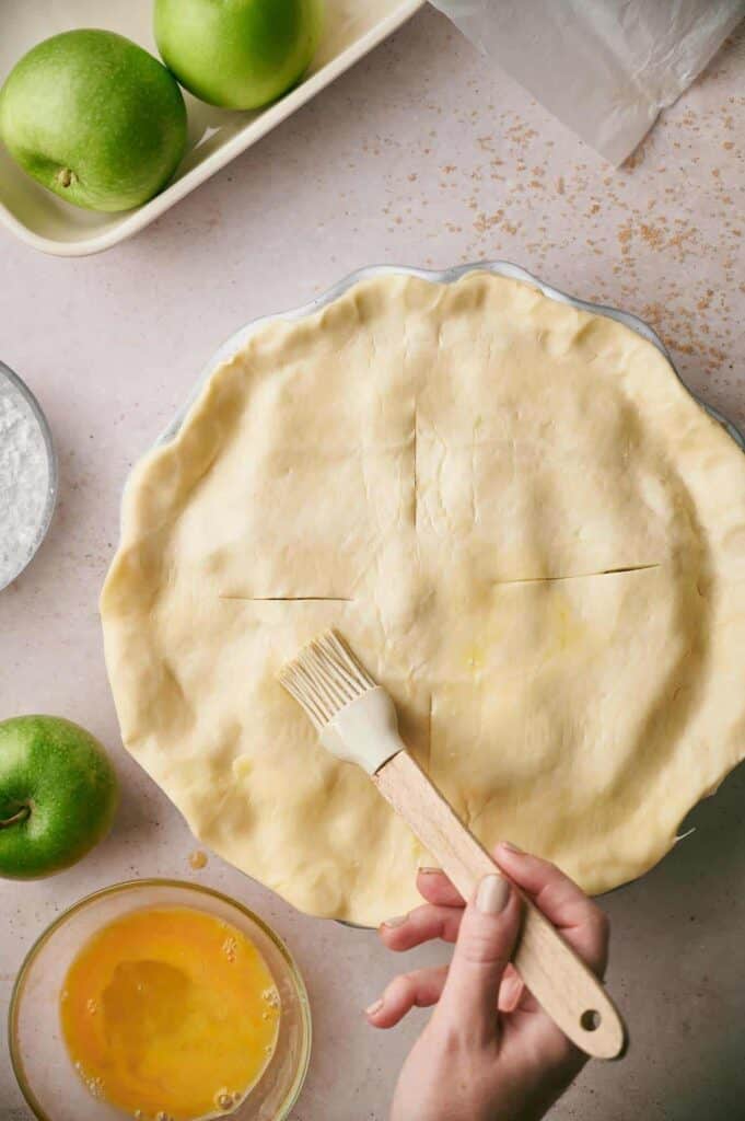 A person is brushing egg wash onto the top crust of an unbaked pie. Nearby, there are green apples, a small bowl of egg wash, and a bowl of powdery substance.