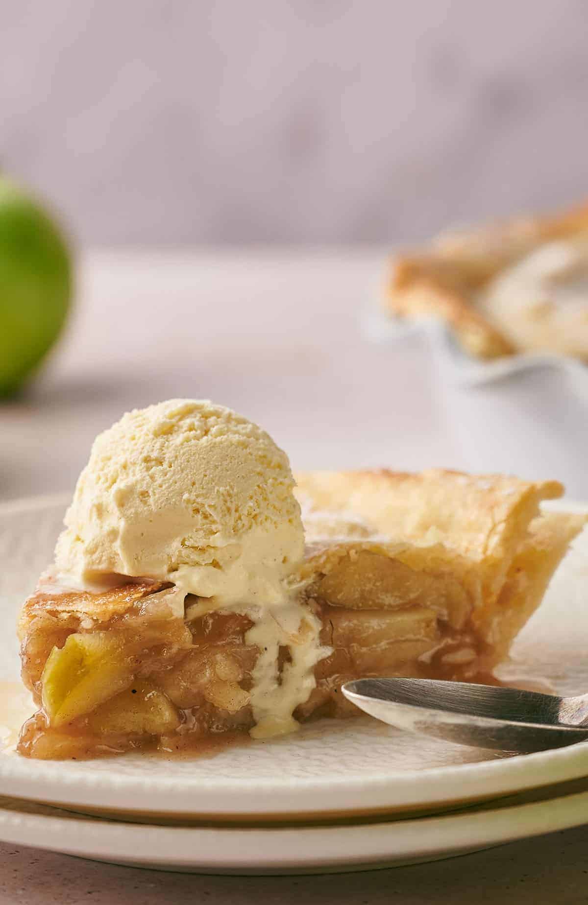 A slice of apple pie topped with a scoop of vanilla ice cream on a plate, with a fork beside it. An out-of-focus green apple and another pie can be seen in the background.