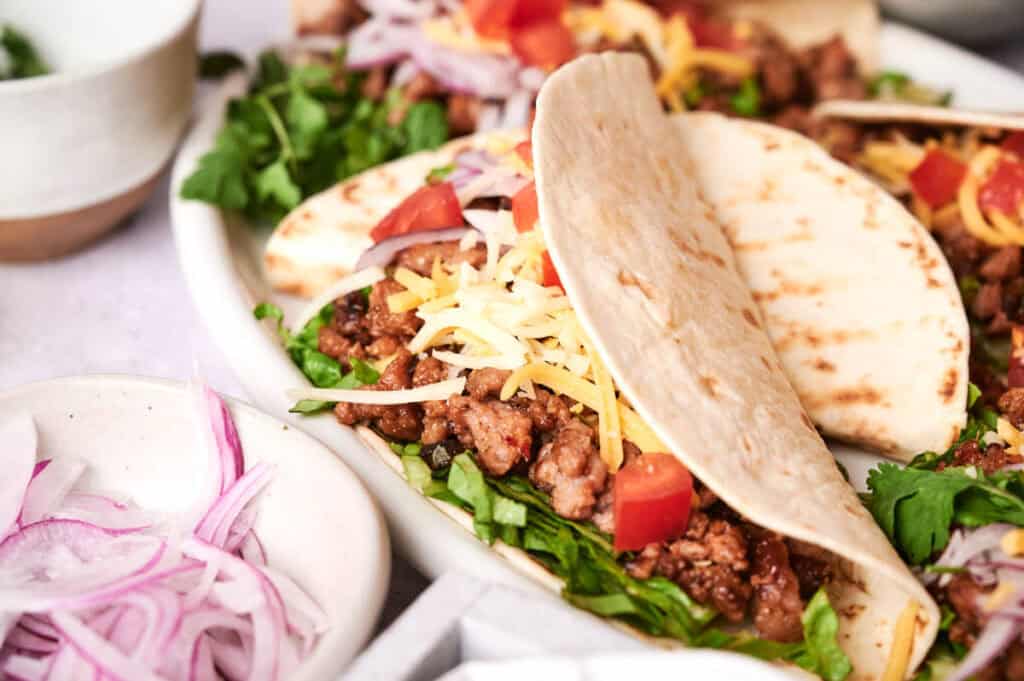 Close-up of a plate with beef tacos filled with ground meat, shredded cheese, lettuce, and chopped tomatoes, garnished with thin slices of red onion. Two small bowls with additional toppings are nearby.