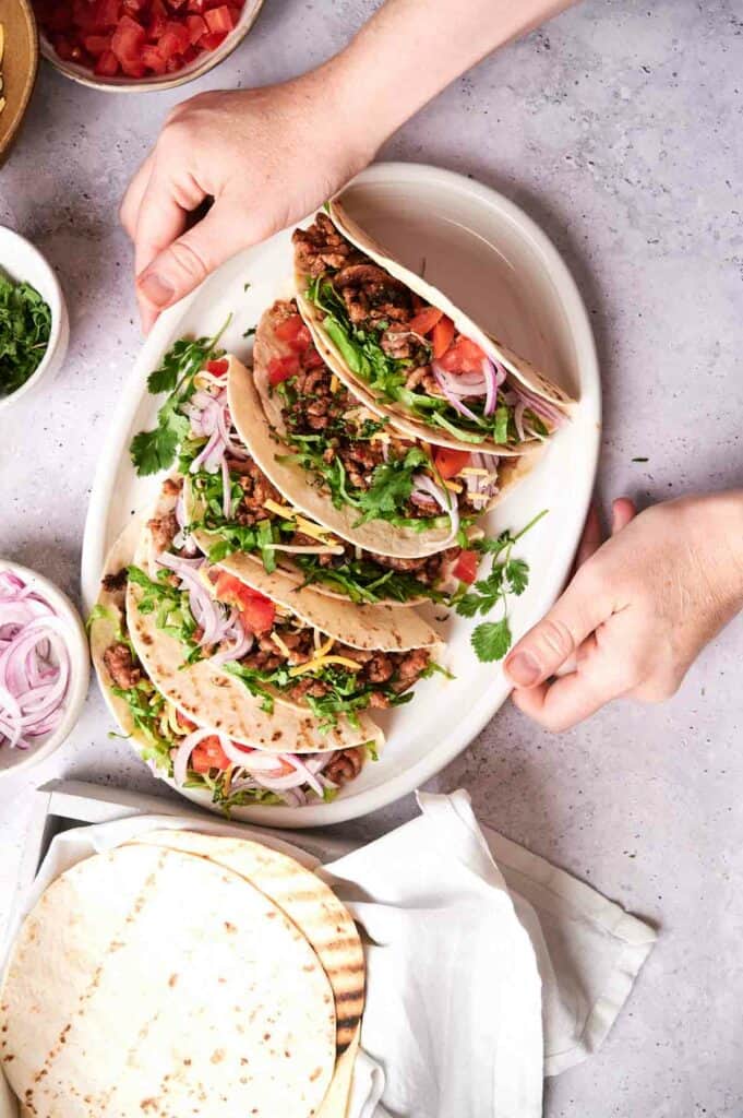 A pair of hands holding a plate with three homemade beef tacos filled with ground meat, tomatoes, red onions, cilantro, and shredded cheese, surrounded by bowls of ingredients and tortillas.