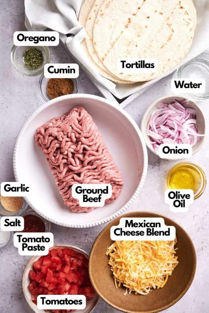 Ingredients for beef tacos arranged on a surface, including tortillas, ground beef, diced tomatoes, tomato paste, cumin, oregano, garlic, salt, sliced onions, olive oil, water, and Mexican cheese blend.