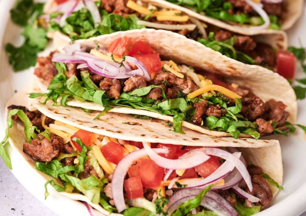 Enjoy our Beef Tacos: three soft tacos filled with seasoned ground beef, shredded lettuce, diced tomatoes, sliced red onions, shredded cheese, and cilantro on a white plate.