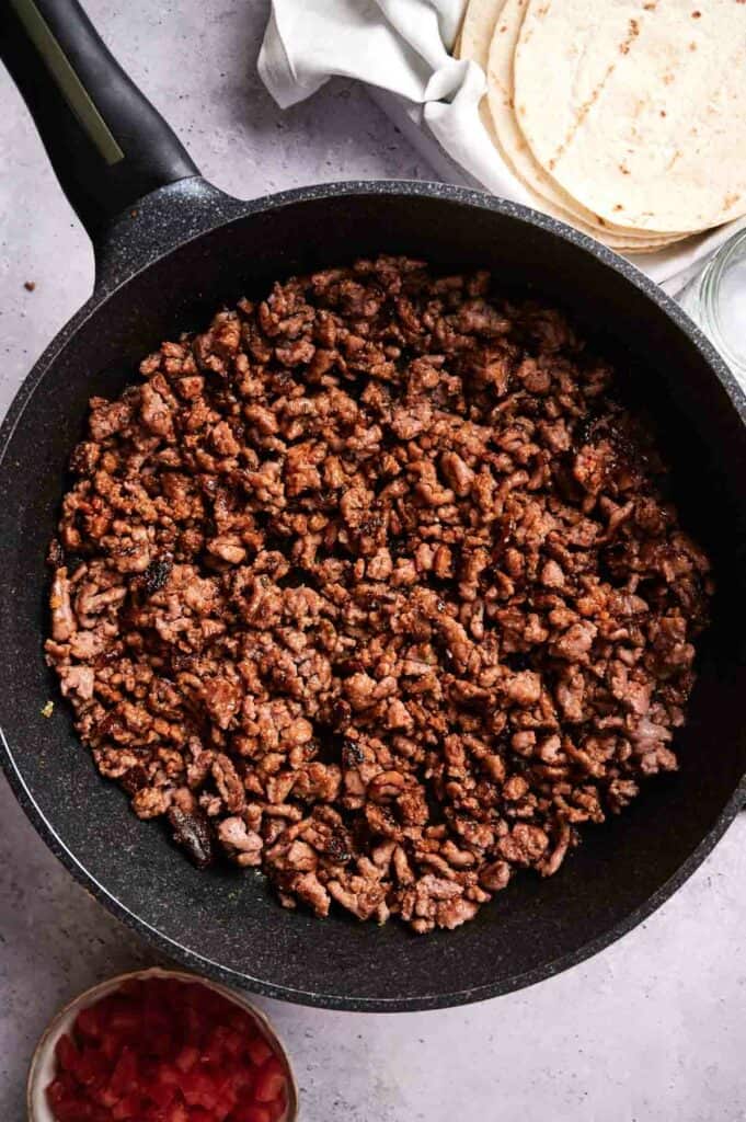 A pan filled with cooked ground beef sits on a countertop, ready for delicious beef tacos. Nearby are tortillas, diced tomatoes in a bowl, and a white cloth.