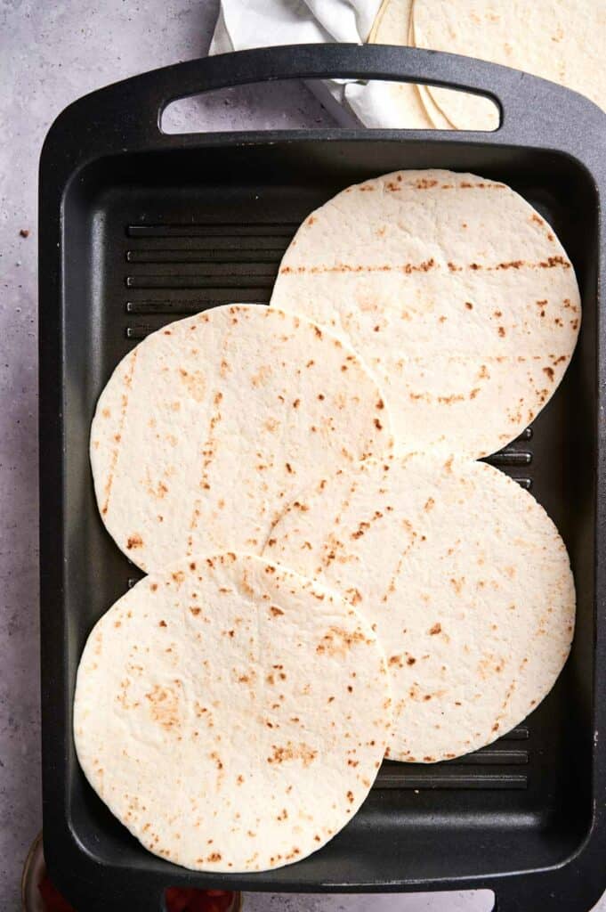Four flour tortillas are arranged on a black griddle, ready to be transformed into delicious beef tacos.