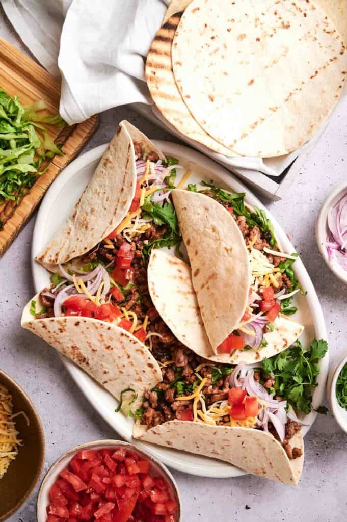 A plate with three juicy beef tacos topped with shredded cheese, tomatoes, and onions. Surrounding the plate are bowls of chopped tomatoes, grated cheese, sliced onions, and a cutting board with shredded lettuce.