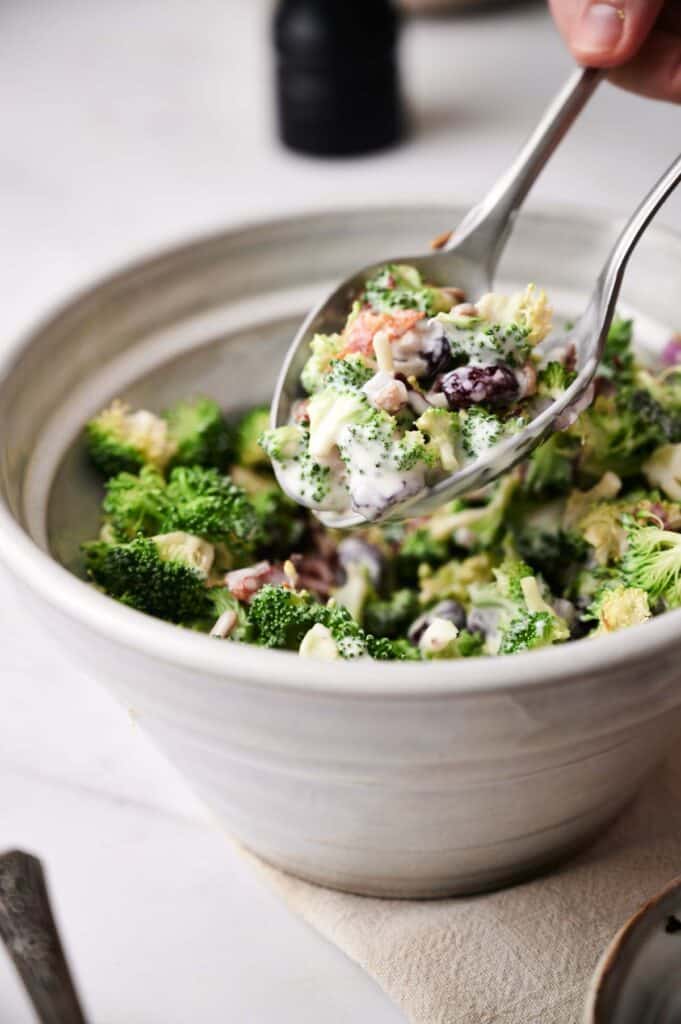 A hand serving a spoonful of broccoli salad with creamy dressing, raisins, and nuts from a white ceramic bowl.