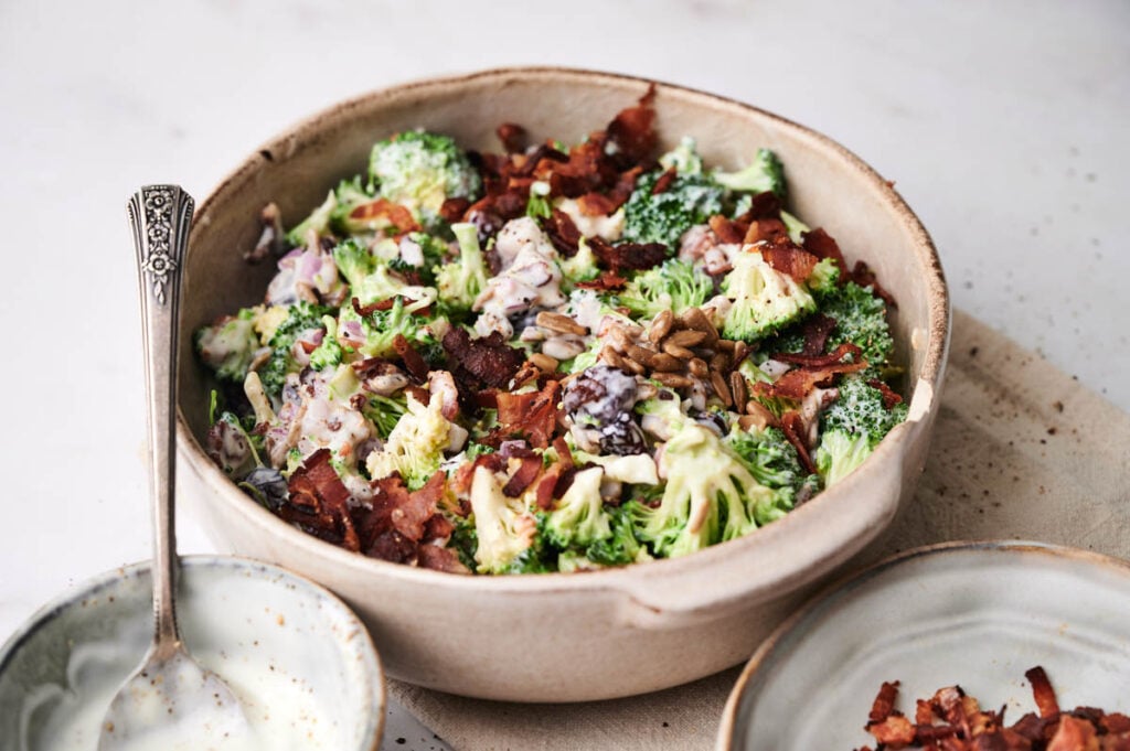 A bowl of broccoli salad with bacon, red onions, sunflower seeds, and creamy dressing sits ready to enjoy, with a spoon and a small bowl of extra dressing beside it.