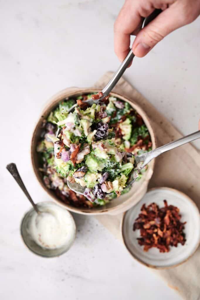 A hand holds serving spoons scooping out a portion of broccoli salad from a bowl. Next to the bowl, there is a small plate of bacon bits and a bowl of creamy dressing, perfect accompaniments for this delicious dish.