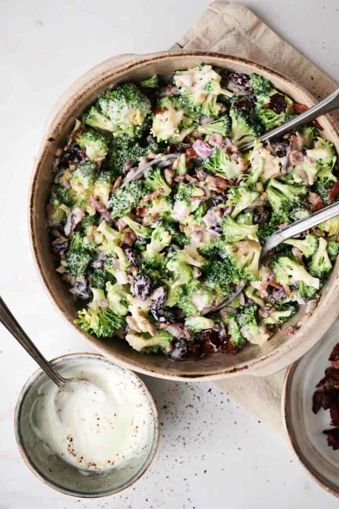 A bowl of broccoli salad with dried cranberries, red onions, and creamy dressing is accompanied by a small bowl of white sauce and a plate of bacon bits on a light-colored surface.