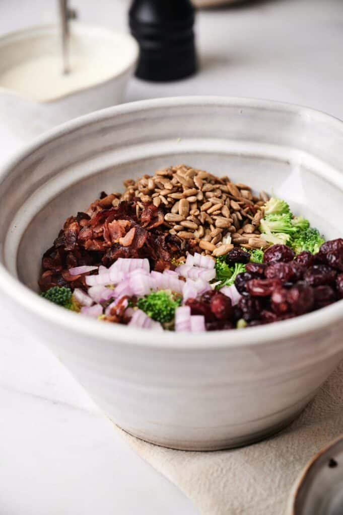 A ceramic bowl filled with ingredients for a delicious broccoli salad, including chopped red onions, dried cranberries, bacon bits, and sunflower seeds.
