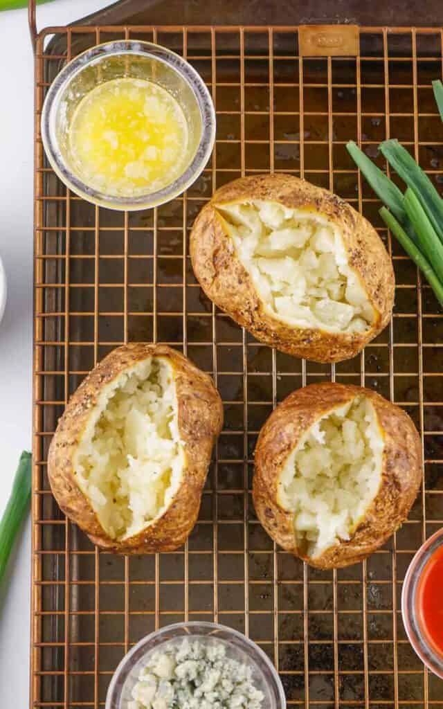 Two Buffalo chicken baked potatoes on a cooling rack with cut green onions, melted butter, and grated cheese in separate containers, viewed from above.