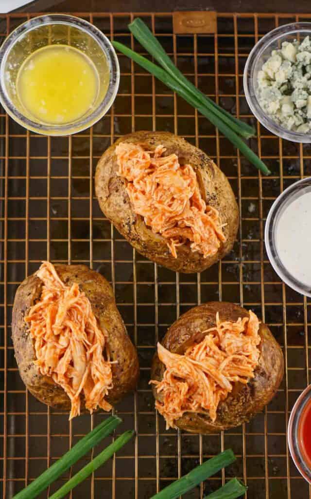 Three Buffalo chicken baked potatoes topped with shredded carrot, displayed on a cooling rack, accompanied by bowls of melted butter and blue cheese dressing.