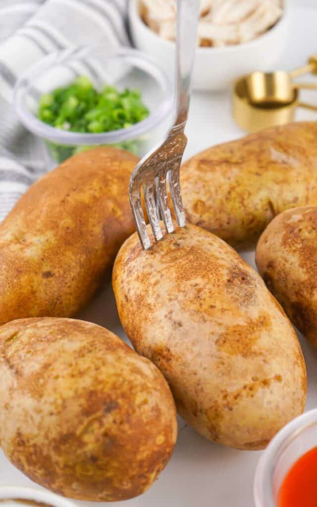 A fork piercing one of several whole, unpeeled Buffalo Chicken Baked Potatoes on a plate, with condiments and chopped green onions in the background.