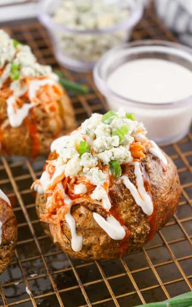 Buffalo Chicken Baked Potatoes topped with blue cheese, diced celery, and drizzled with ranch dressing, served on a wire rack.