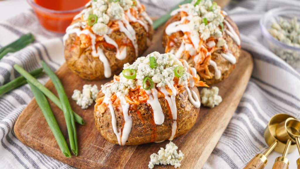 Buffalo Chicken Baked Potatoes topped with blue cheese and ranch dressing, served on a wooden board with green onions and a side sauce.