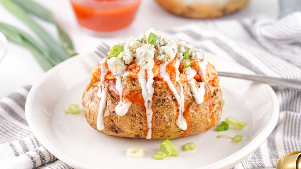 Buffalo Chicken Baked Potatoes: Baked potato topped with blue cheese crumbles, ranch, hot sauce, and chopped green onions, served on a white plate with a fork.