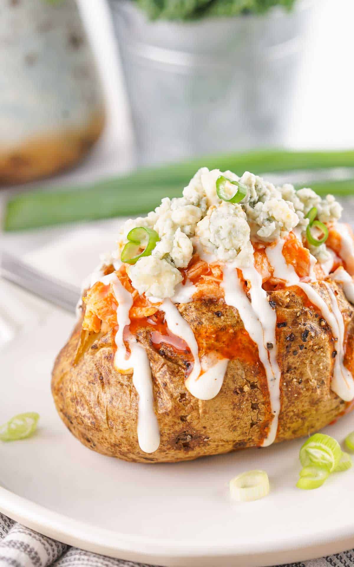 Buffalo Chicken Baked Potatoes topped with blue cheese and drizzled with ranch dressing, garnished with sliced green onions on a white plate.