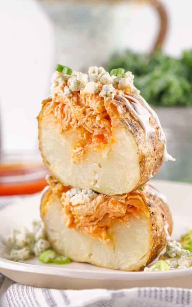 Buffalo Chicken Baked Potatoes: A baked potato sliced open and filled with buffalo chicken, topped with blue cheese crumbles and chopped green onions, served on a white plate.