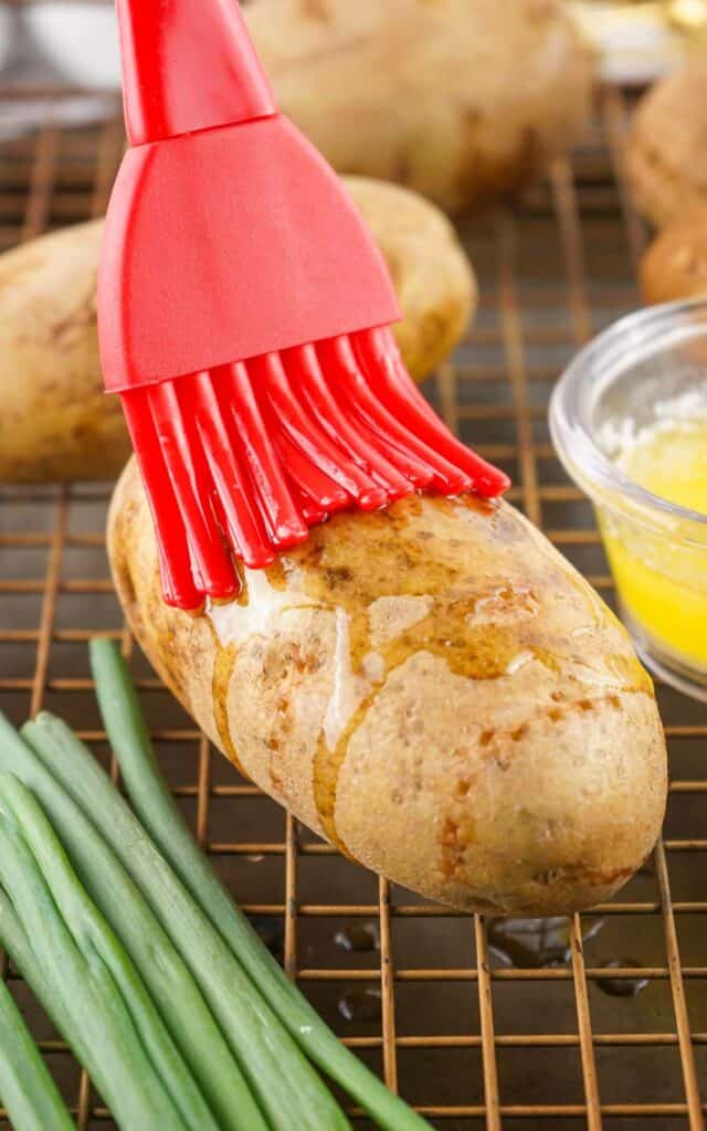 A red brush applying butter to Buffalo Chicken Baked Potatoes on a wire rack, with green onions and a bowl of melted butter nearby.