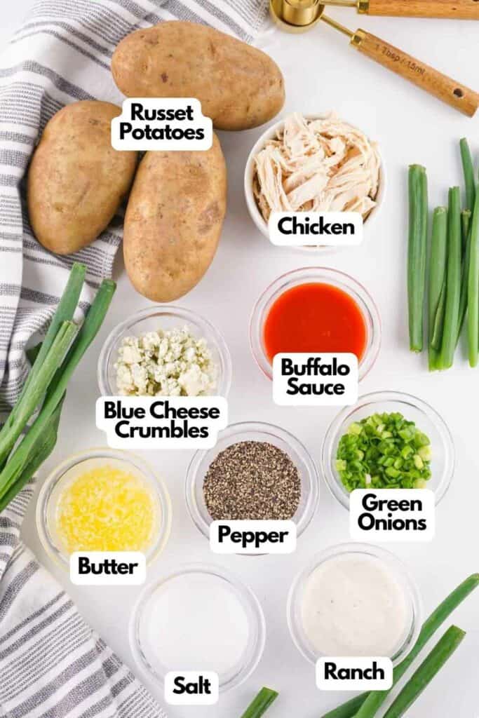 Overhead view of ingredients for Buffalo Chicken Baked Potatoes on a white surface, including russet potatoes, shredded chicken, buffalo sauce, green onions, blue cheese, and seasonings.
