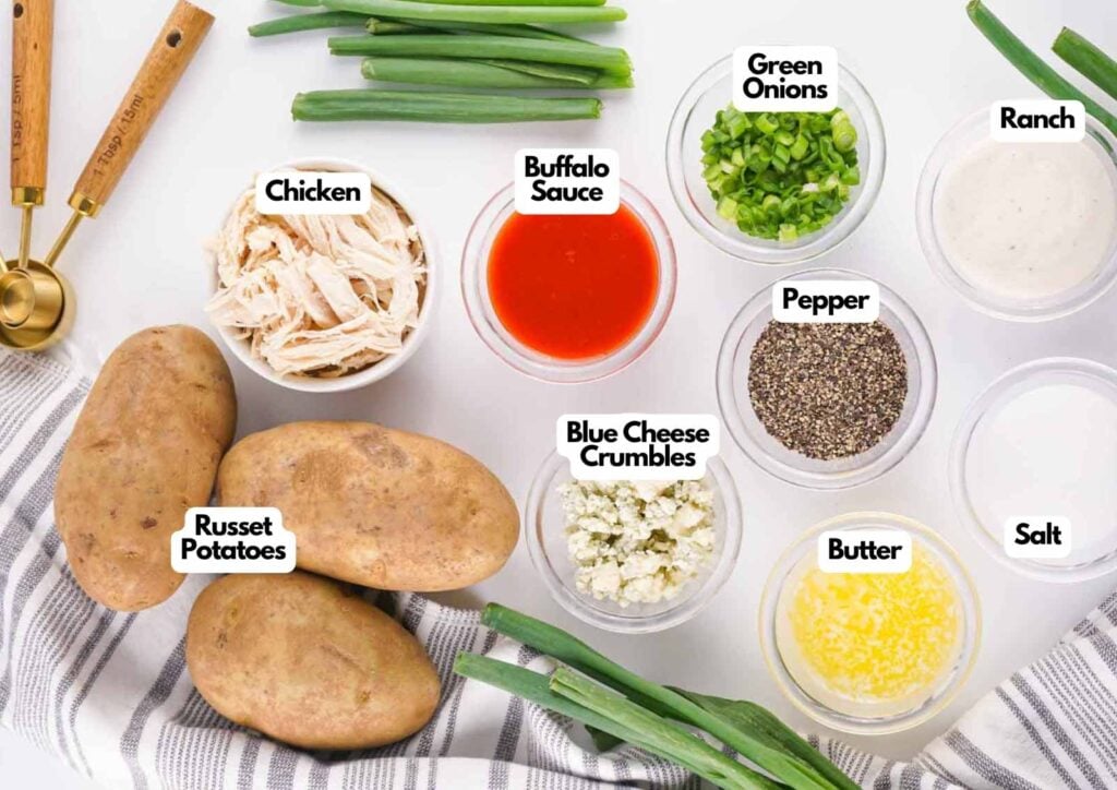 Top-down view of labeled ingredients for Buffalo Chicken Baked Potatoes, including russet potatoes, shredded chicken, buffalo sauce, blue cheese crumbles, green onions, ranch, pepper, butter, and salt.