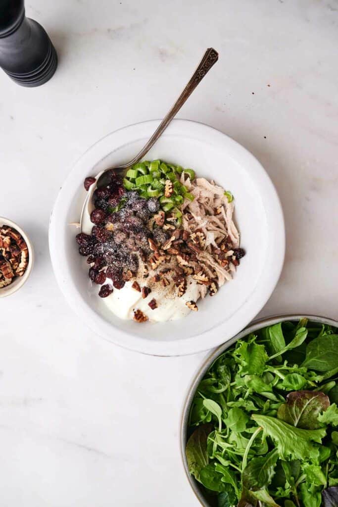 A bowl brimming with chicken salad ingredients, including shredded chicken, celery, cranberries, and chopped nuts, is next to a plate of leafy greens.
