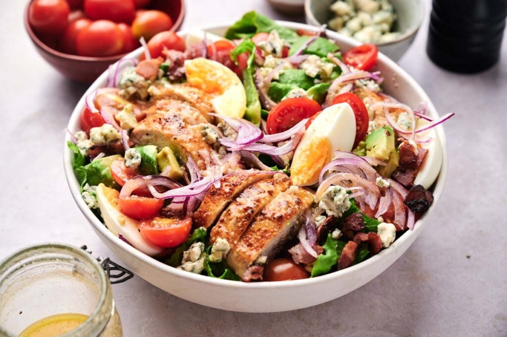 A classic cobb salad featuring sliced grilled chicken, halved cherry tomatoes, hard-boiled eggs, red onions, bacon bits, avocado, blue cheese crumbles, and fresh leafy greens.