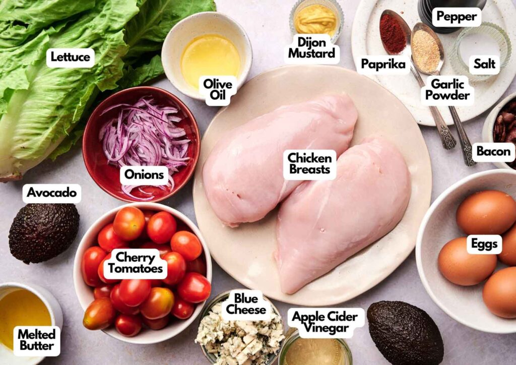 Top-down view of labeled cobb salad ingredients, including chicken breasts, eggs, bacon, veggies, blue cheese, and various seasonings neatly arranged on a table.