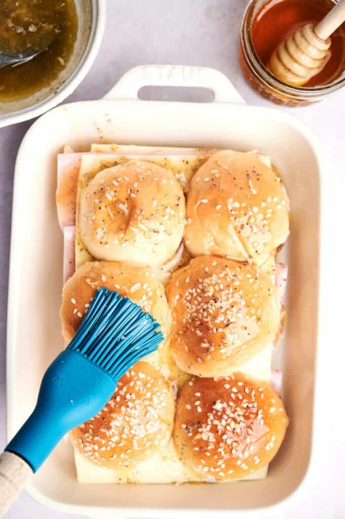 A baking dish with six Ham and Cheese Sliders being brushed with butter. Honey and a basting brush are visible nearby. The buns are sprinkled with salt and look freshly baked.