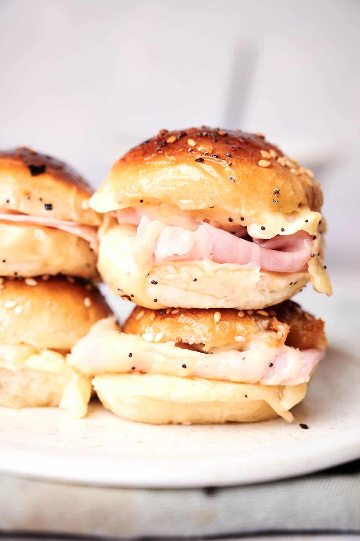 Three delectable Ham and Cheese Sliders with sesame seed toppings are stacked on a pristine white plate.