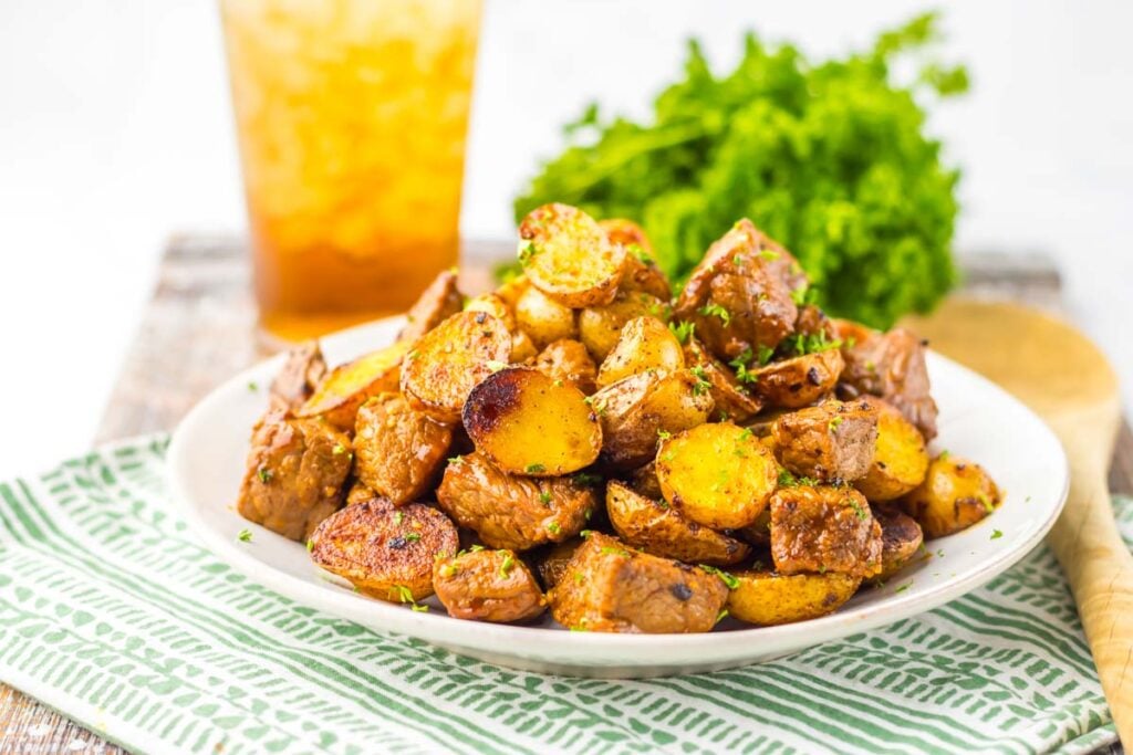 A plate of Honey Chipotle Steak and Potatoes, garnished with fresh herbs, sits on a patterned napkin, with a glass of iced tea and leafy greens in the background.