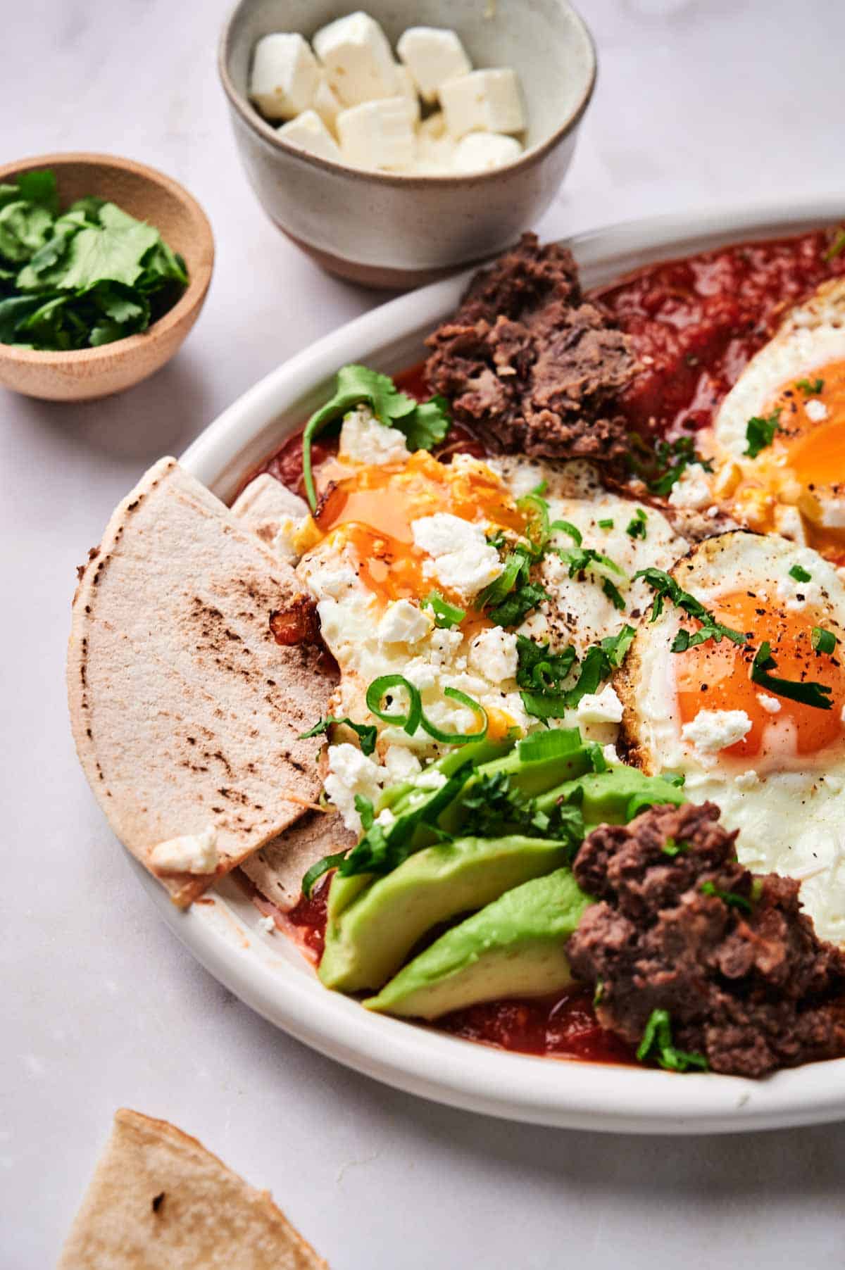 A plate containing huevos rancheros with fried eggs, sliced avocado, refried beans, and tortilla slices. Small bowls of chopped herbs and diced cheese are in the background. The classic breakfast dish makes for a satisfying meal rich in flavor and texture.
