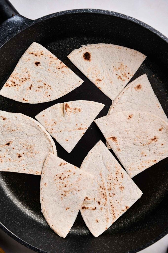 Several pieces of cut flour tortillas, perfect for huevos rancheros, are toasting in a black frying pan.