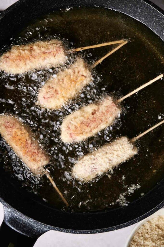 Five breaded skewers, reminiscent of Korean Corn Dogs, are frying in hot oil in a pan.