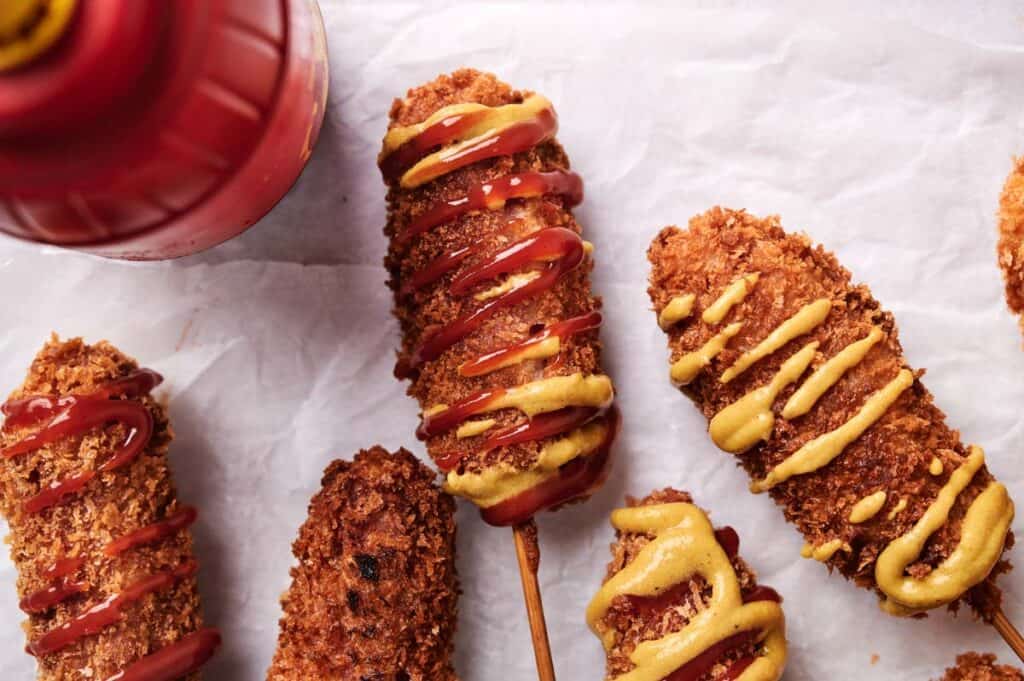 Close-up of five Korean corn dogs with crispy coatings placed on parchment paper. Some are topped with mustard, and some with ketchup. A red bottle, presumably ketchup, is visible on the left.