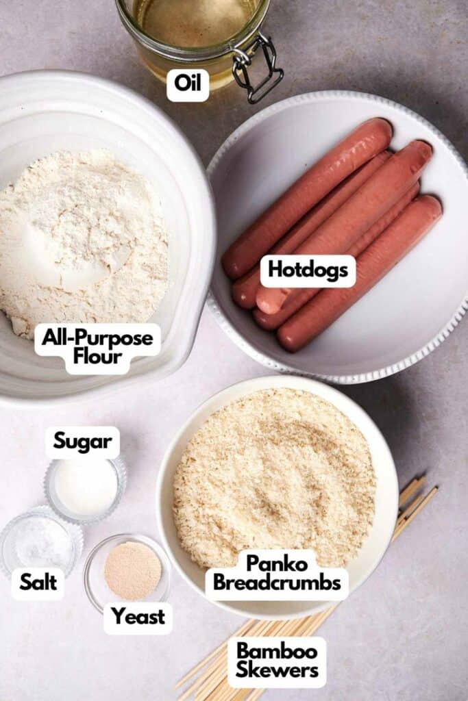 Top view of ingredients for making Korean Corn Dogs: bowls of all-purpose flour, sugar, salt, yeast, panko breadcrumbs, four hotdogs, a jar of oil, and bamboo skewers.