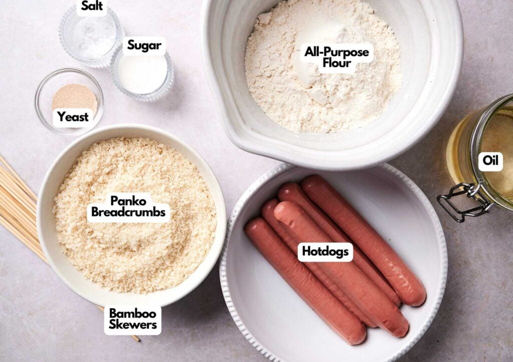 Bowls containing all-purpose flour, panko breadcrumbs, hotdogs, and ingredients like yeast, salt, sugar, oil, and bamboo skewers—perfect essentials for making delicious Korean Corn Dogs—are arranged on a countertop.