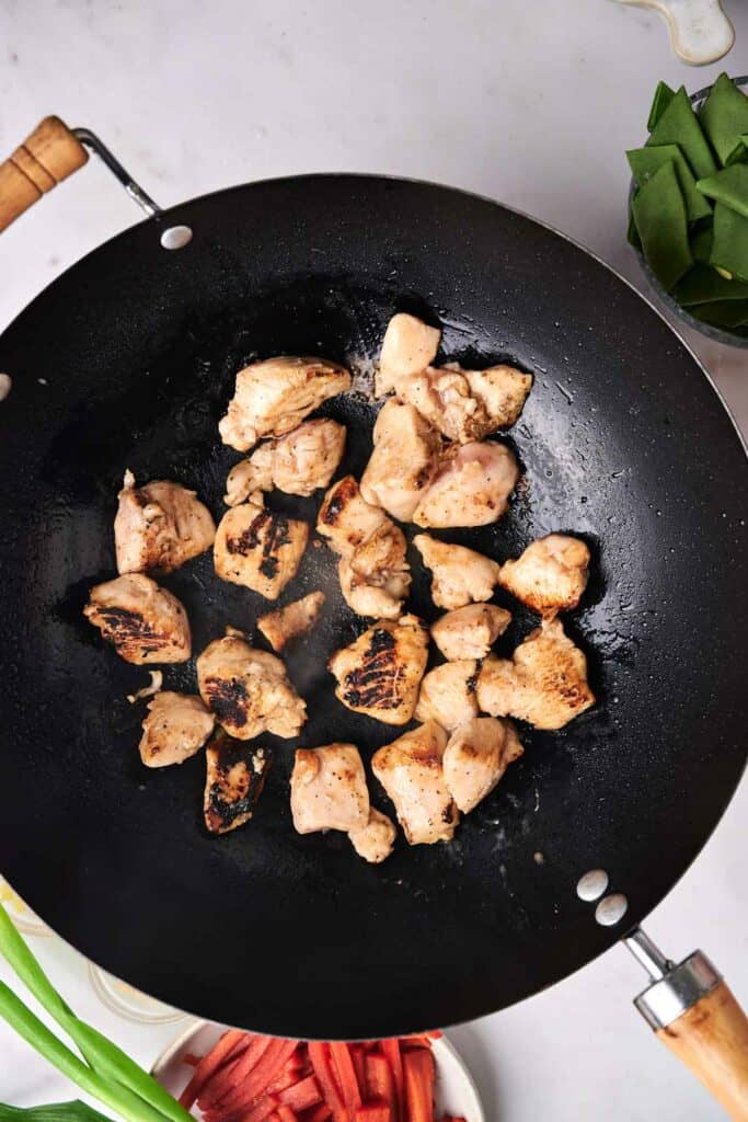 Chunks of chicken being cooked in a black wok on a stove, sizzling away to create an irresistible kung pao chicken. Surrounding the wok are bowls of sliced vegetables and greens.