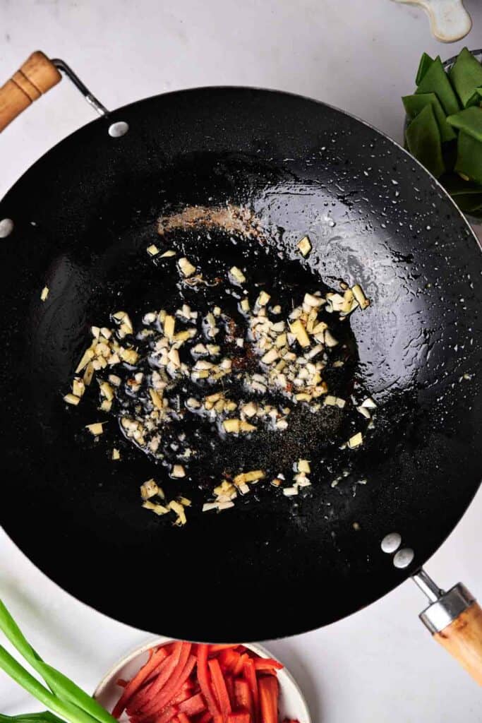 Chopped garlic being saut&eacute;ed in a black wok, setting the stage for a delicious kung pao chicken. Green onions and sliced red bell peppers are placed nearby, ready to join the savory mix.