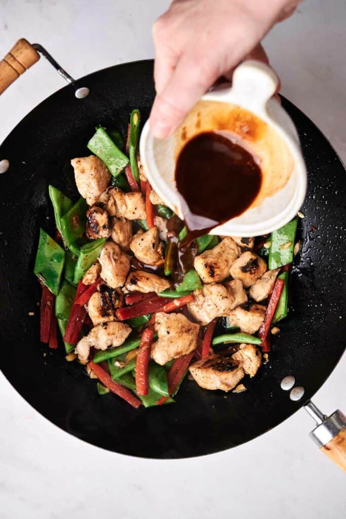 A hand pouring sauce from a white pitcher into a wok containing stir-fried chicken chunks, green snap peas, and red bell pepper strips, creating a delicious twist on kung pao chicken.