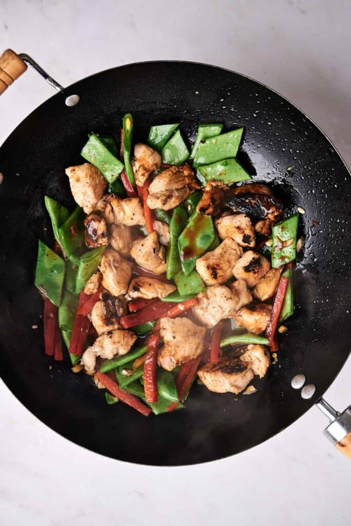 A wok containing cooked chicken pieces, snap peas, and red bell pepper strips is on a white surface, reminiscent of a delicious kung pao chicken.